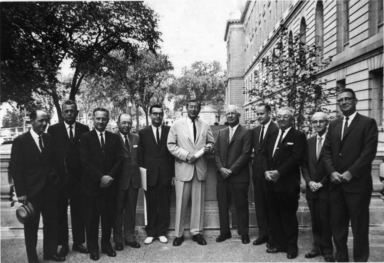 A group of eleven men, dressed in formal suits, pose together outdoors in front of a historic building.