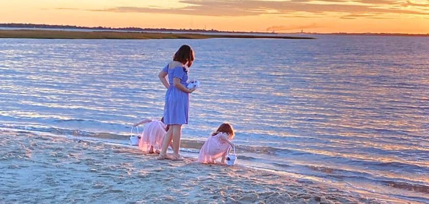 Two children play by the water's edge at sunset, with one standing and the other crouching by the shore.