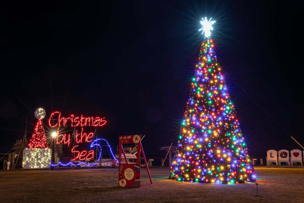 A brightly lit Christmas tree and festive lights display saying "Christmas by the Sea" at night.