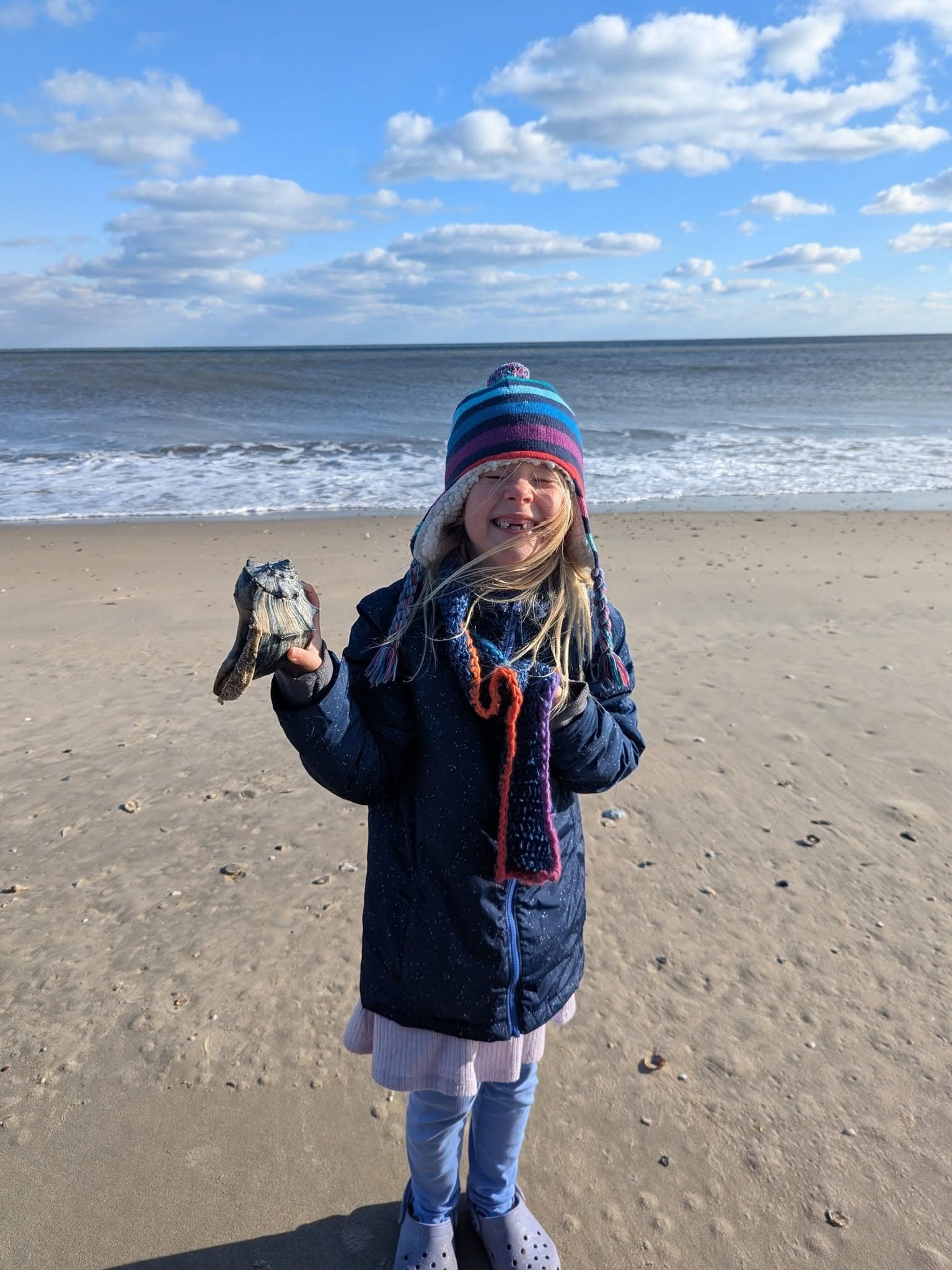 A smiling girl in a winter coat holds a seashell while standing on a beach with a blue sky and ocean in the background.
