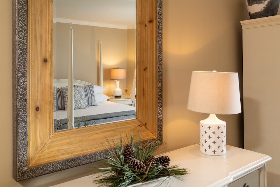 A cozy bedroom is reflected in a wooden-framed mirror, featuring a lamp and decorative pinecones on a bedside table.