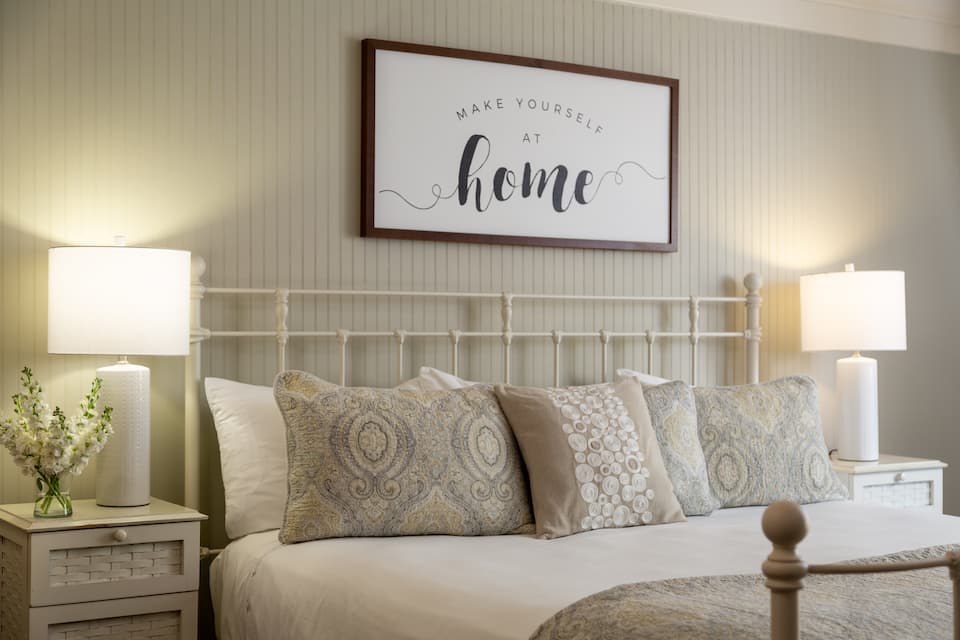 Cozy bedroom featuring a bed with decorative pillows, two bedside lamps, and a wall sign that reads "Make Yourself at Home."