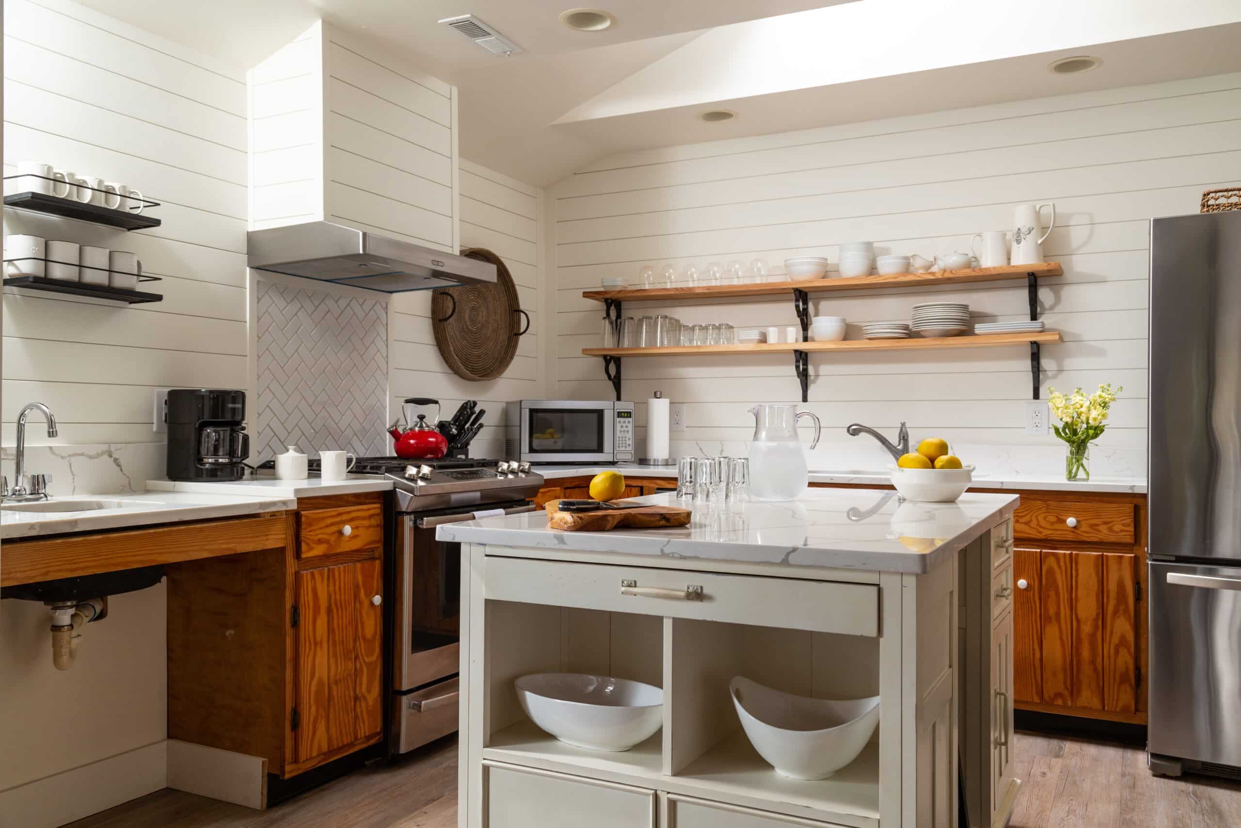 A bright, modern kitchen featuring wooden cabinets, a stainless steel refrigerator, and open shelving with dishware.