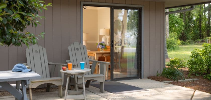 A cozy outdoor seating area with two adirondack chairs, a small table, and a view into a well-lit room through sliding glass doors.