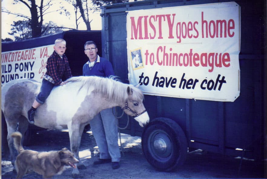 Carlton Leonard rides Misty of Chincoteague beside his father Don Leonard, with a horse trailer bearing a sign that reads "MISTY goes home to Chincoteague to have her colt."