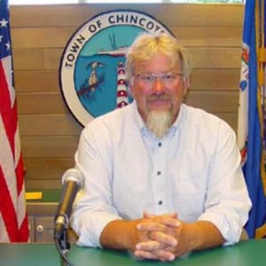 A man with glasses and a beard sits at a table in front of a town seal and flags.