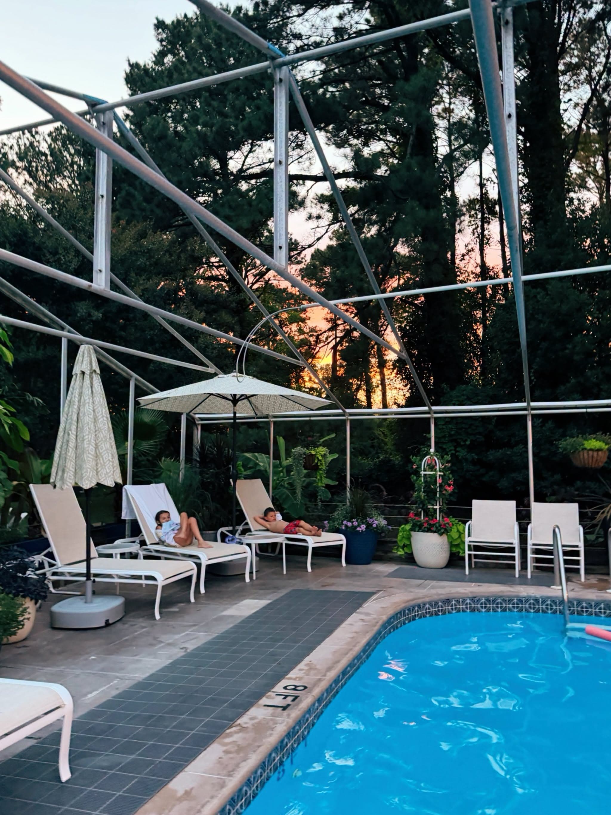 A serene poolside scene featuring loungers, an umbrella, and lush greenery at sunset.