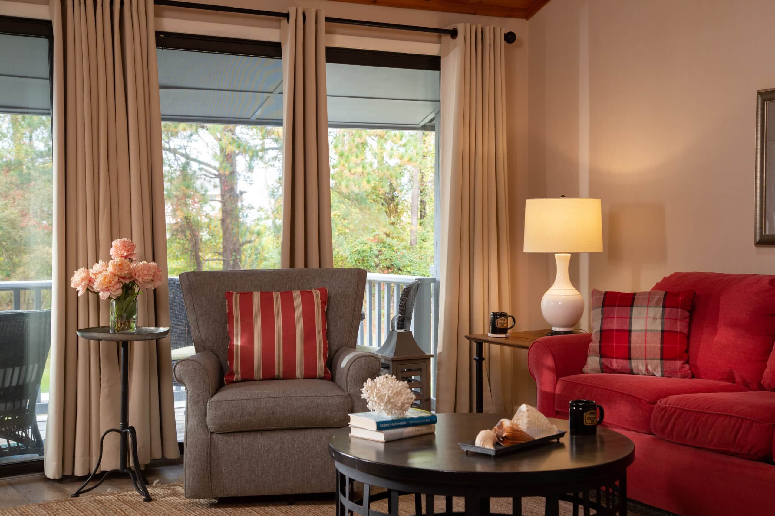 A cozy living room featuring a gray armchair with a striped cushion, a red sofa, a round coffee table, and large windows with sheer curtains revealing a view of greenery outside.