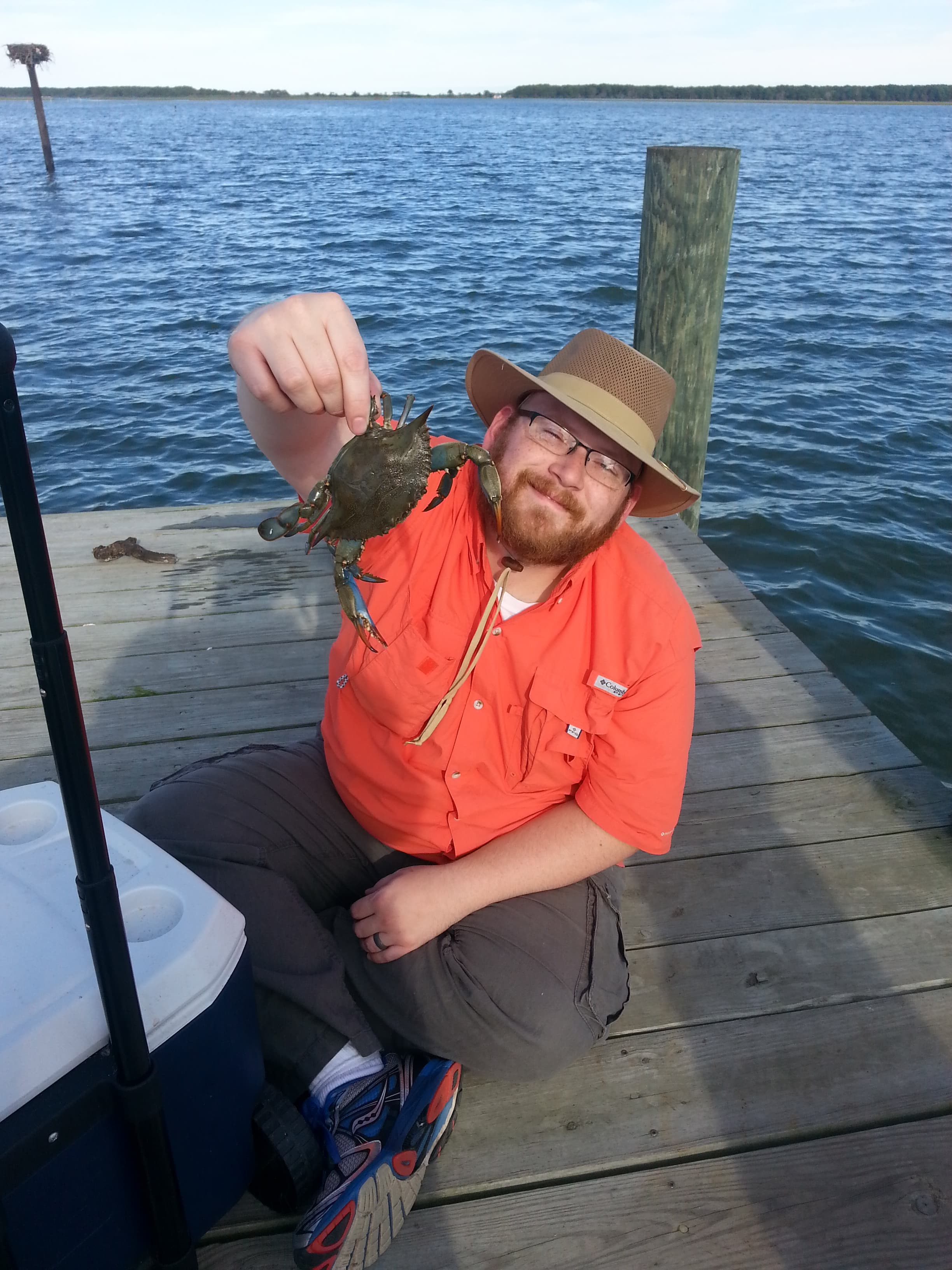 A man in an orange shirt and hat holds up a crab while sitting on a wooden dock by a body of water.
