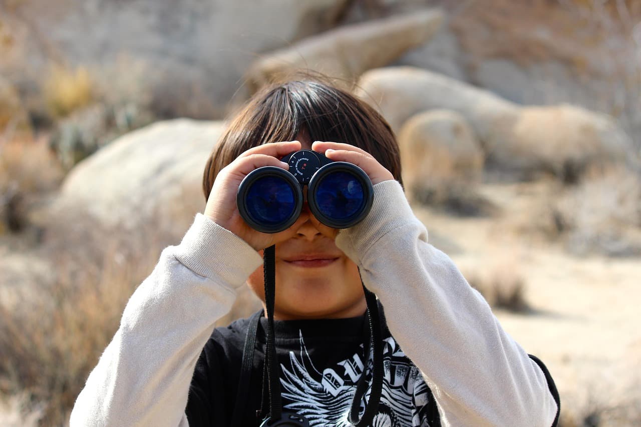 A child holds binoculars up to their eyes, smiling in a rocky outdoor setting. A child holds binoculars up to their eyes, smiling in a rocky outdoor setting.