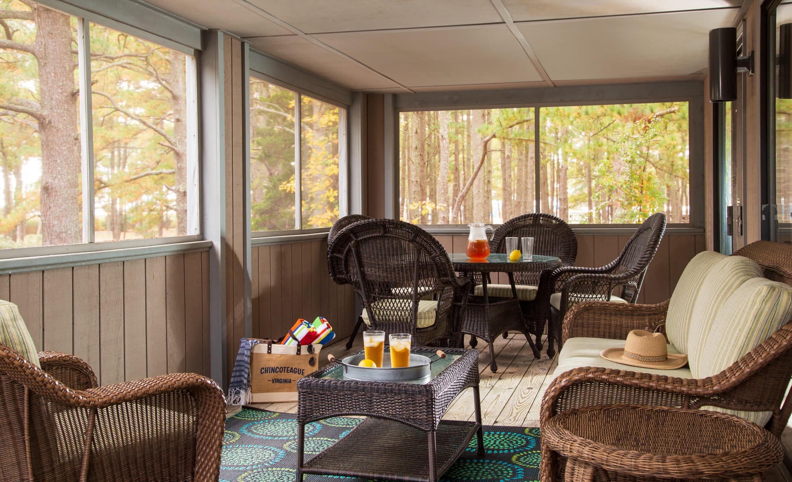 A cozy screened porch with wicker furniture, a coffee table, and drinks, surrounded by trees.