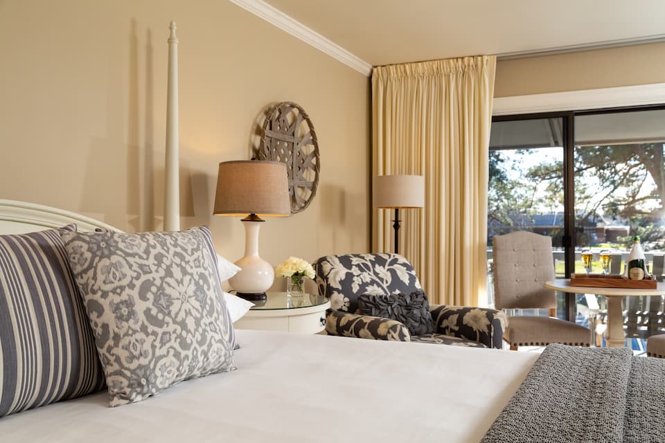 A cozy hotel room featuring a bed with decorative pillows, a chair, a lamp, and large windows with natural light.