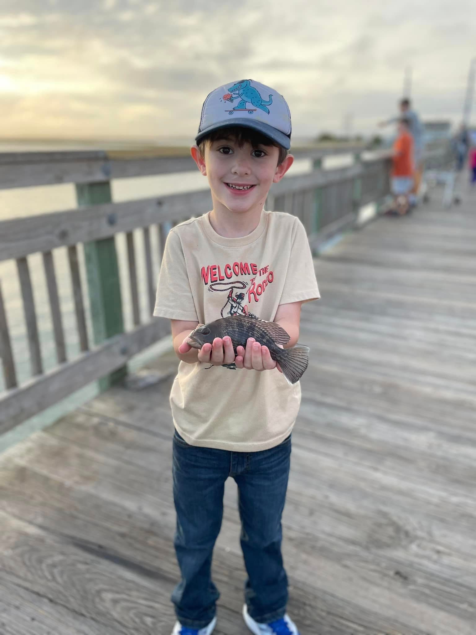 A smiling boy holding a fish on a wooden pier at sunset.