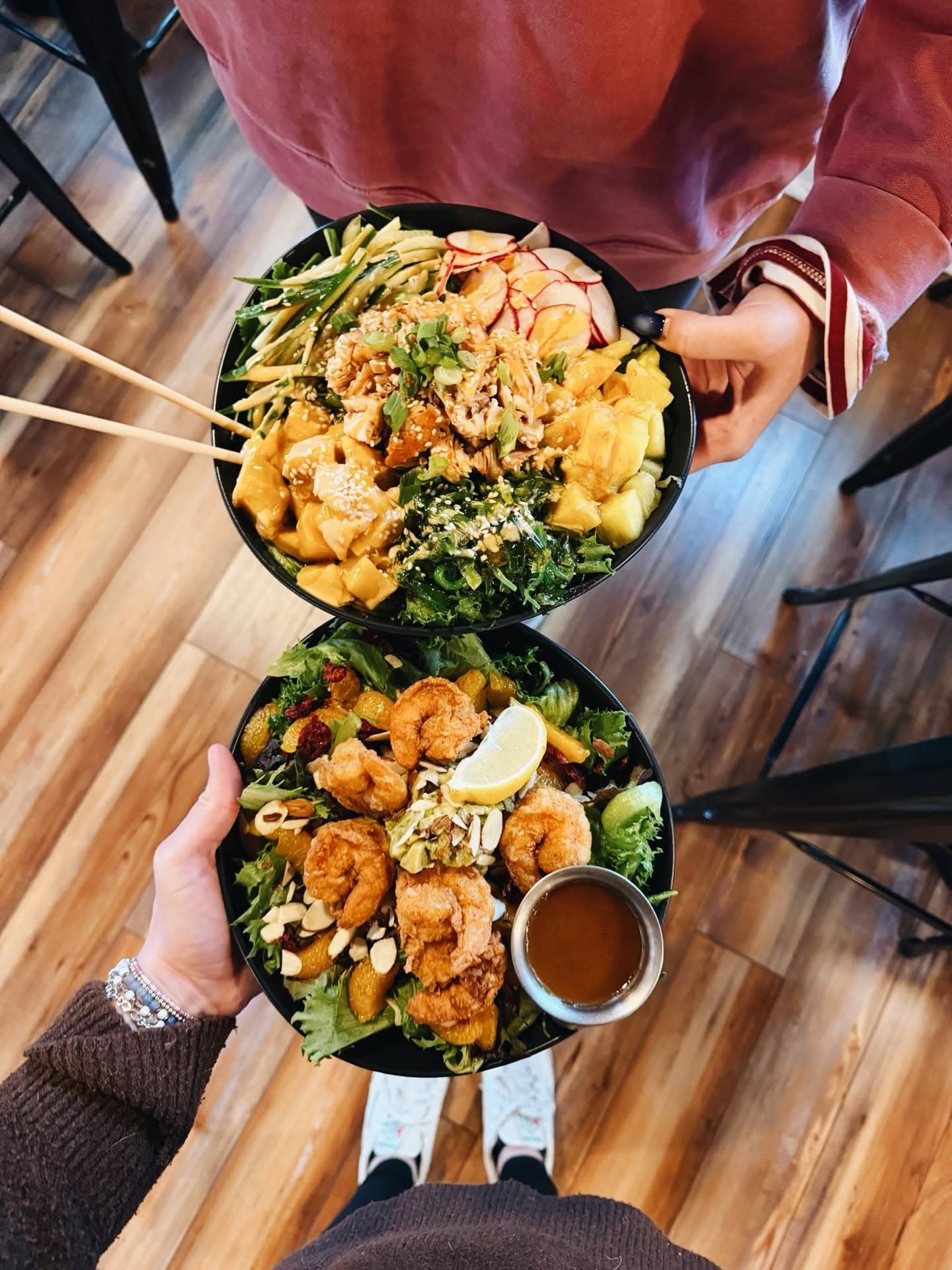 Two colorful salad bowls topped with shrimp and various fresh ingredients are held by a person in a cozy setting.