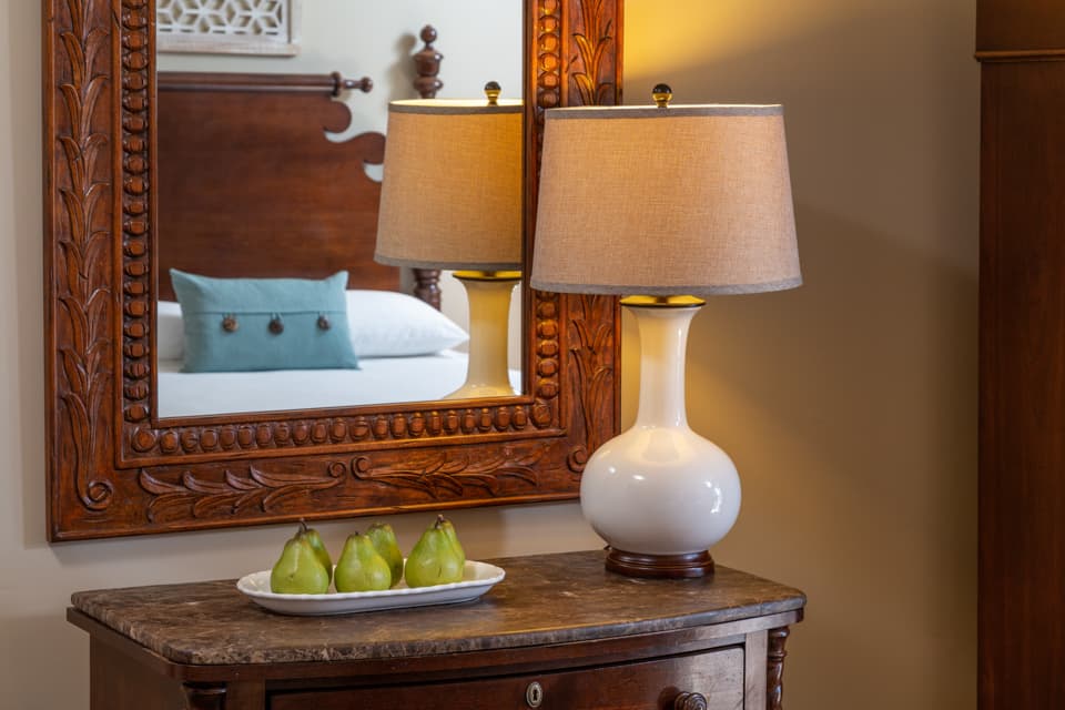 A stylish table lamp sits next to a bowl of pears, reflecting in a decorative mirror above an ornate wooden dresser.