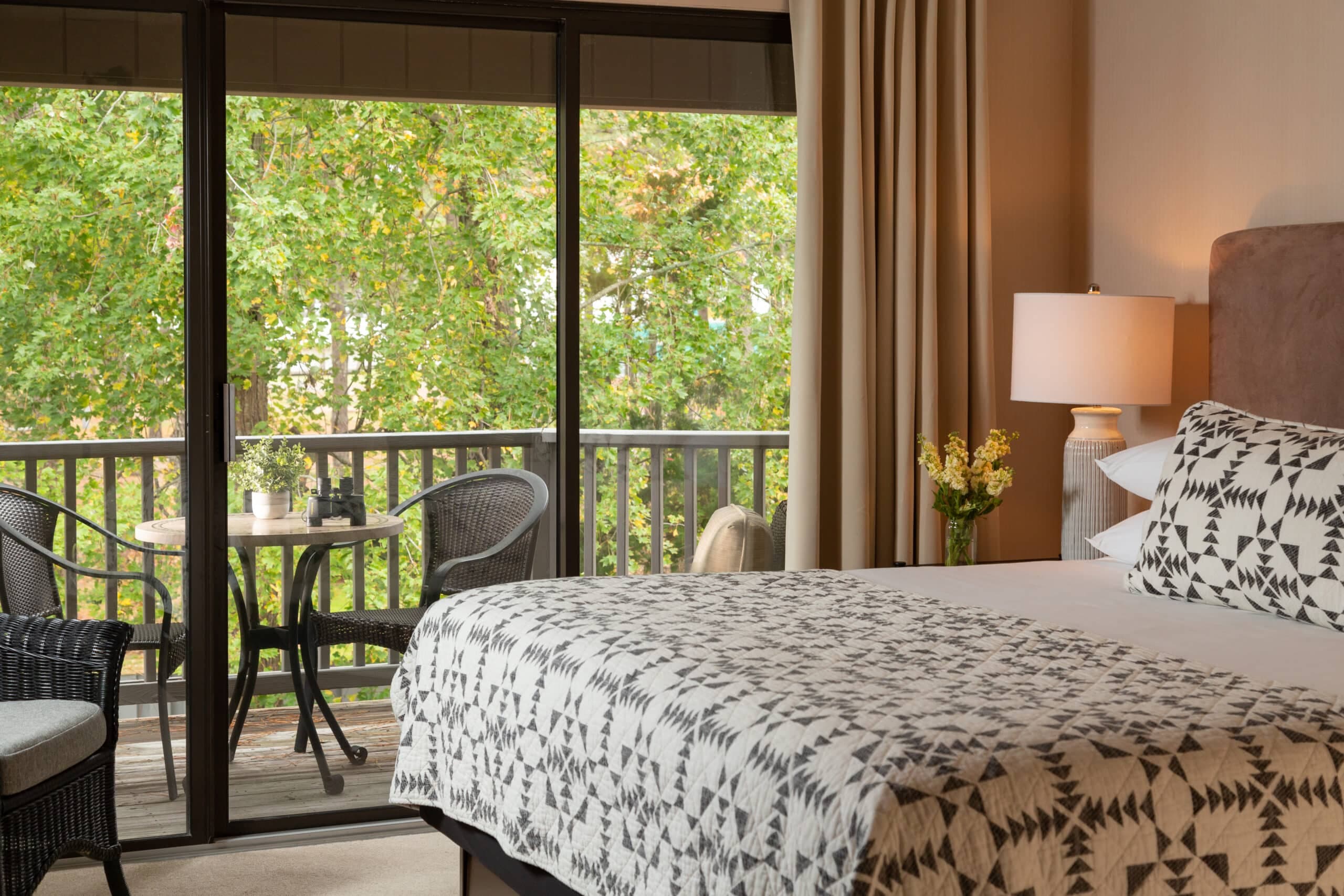 Cozy bedroom with a patterned bedspread, a lamp, and a balcony view featuring greenery.