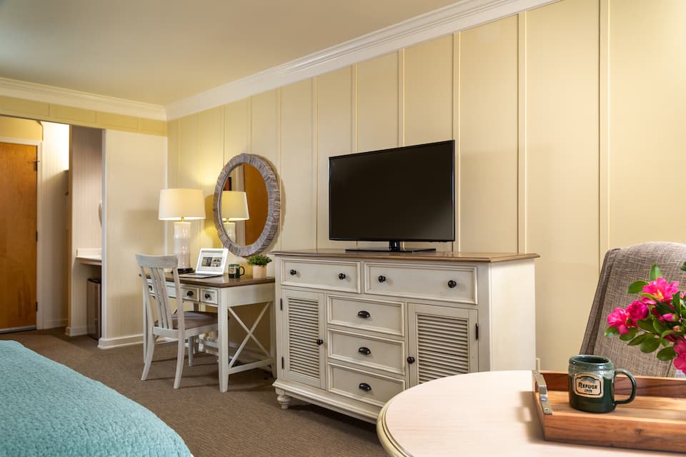 A cozy hotel room featuring a bed, television, dresser, desk, and flower arrangement.