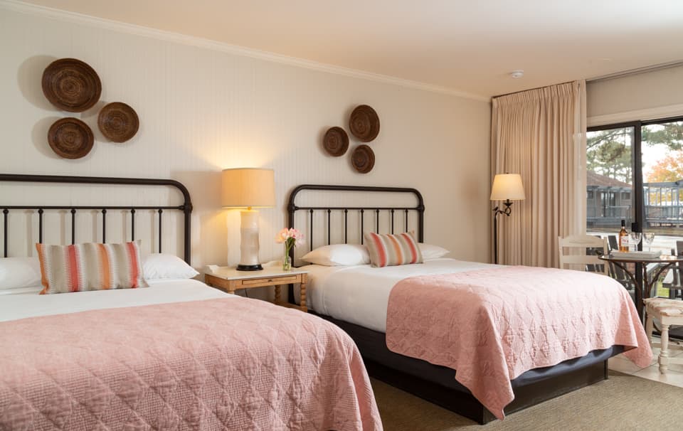 A cozy hotel room with two beds, pastel pink quilts, a lamp, and a small dining table near a window.