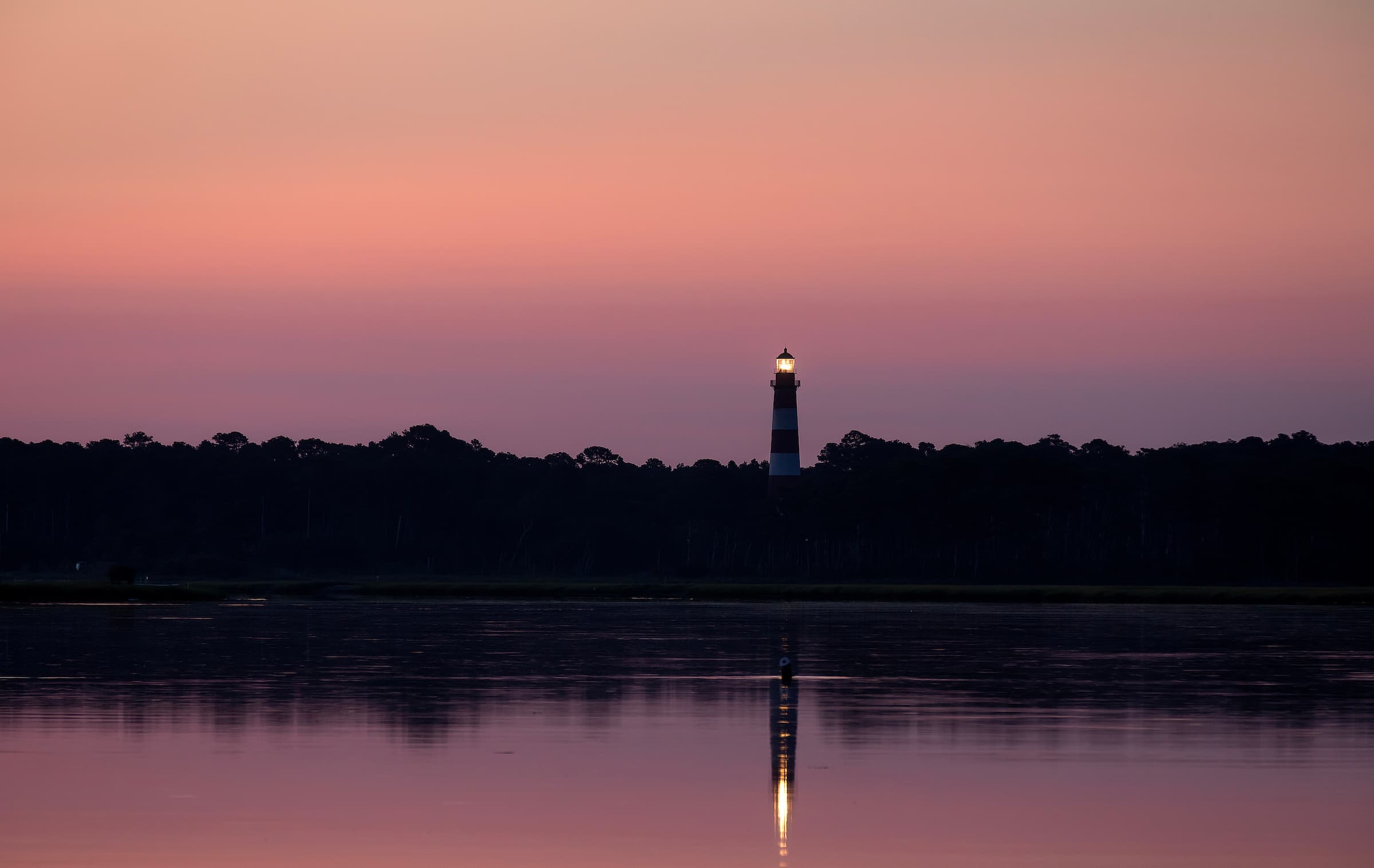 A lighthouse stands against a pastel sunset, reflecting in calm waters.