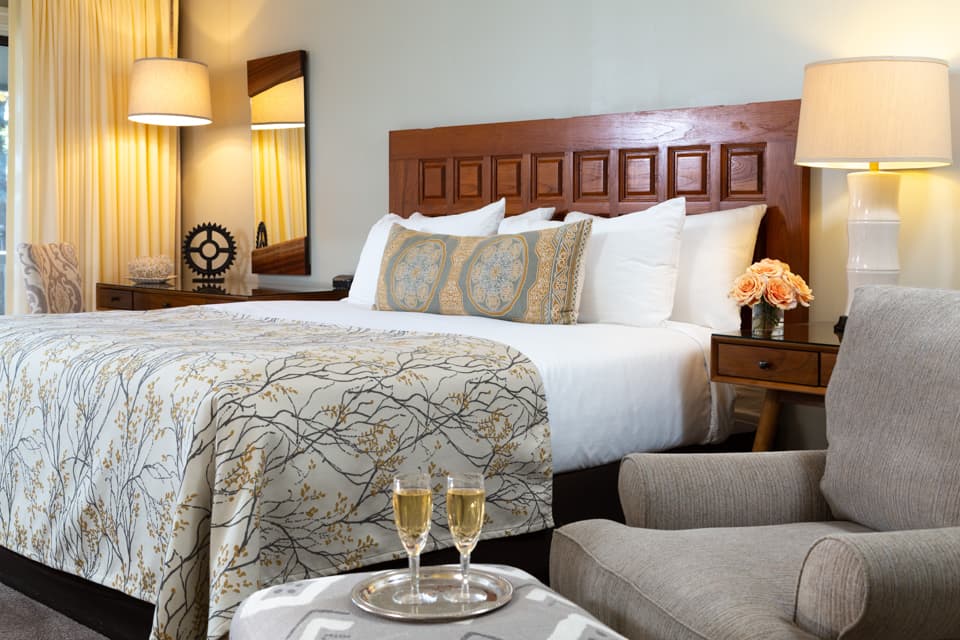 A cozy hotel room featuring a neatly made bed with decorative pillows, a sitting area, and two glasses of champagne on a tray.
