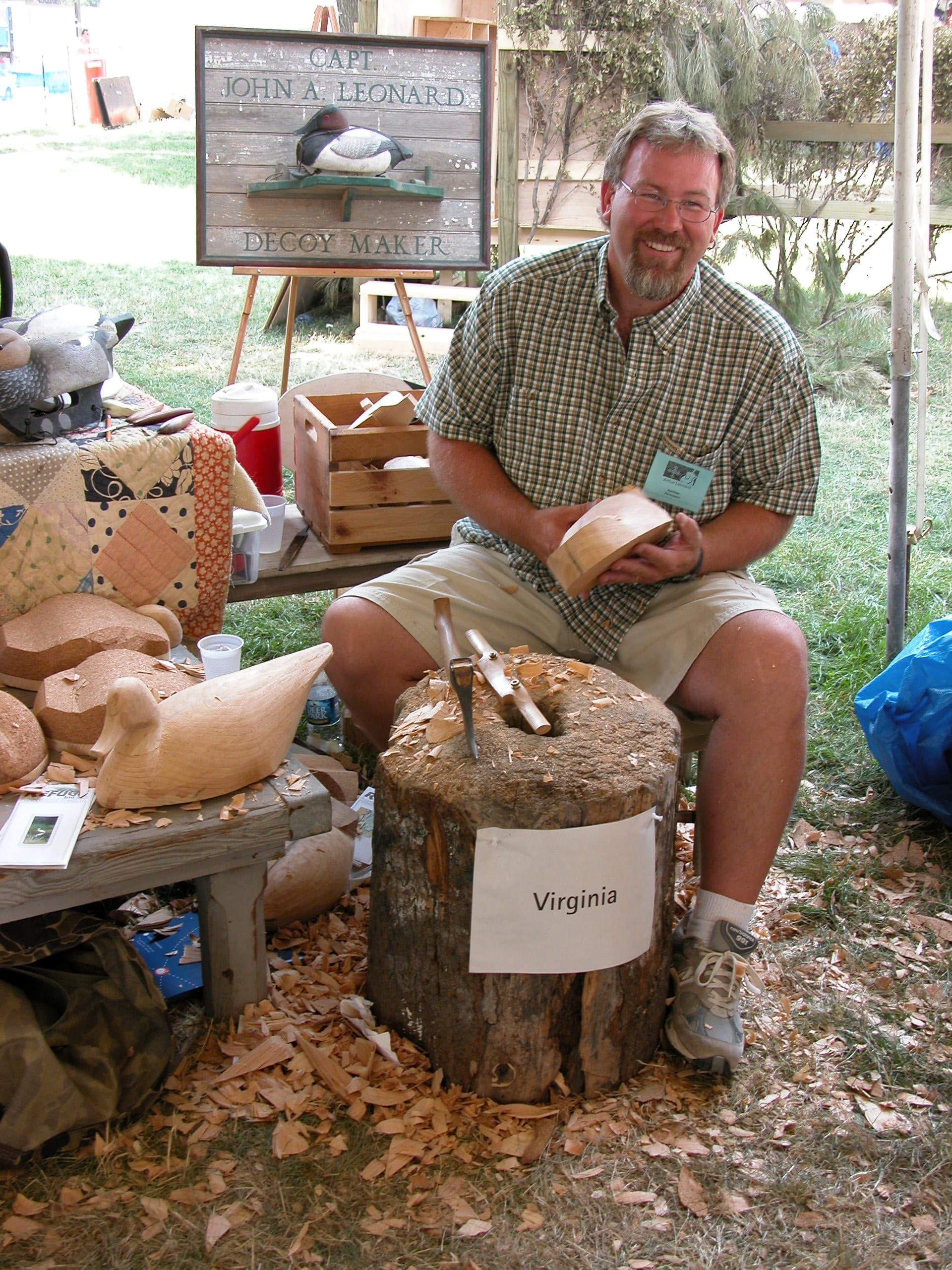 Capt. Arthur Leonard carving a decoy at the Smithsonian Folklife Festival on the National Mall, seated on a stump with shavings around him, roughed-out bodies on the bench and his Decoy Maker sign behind.