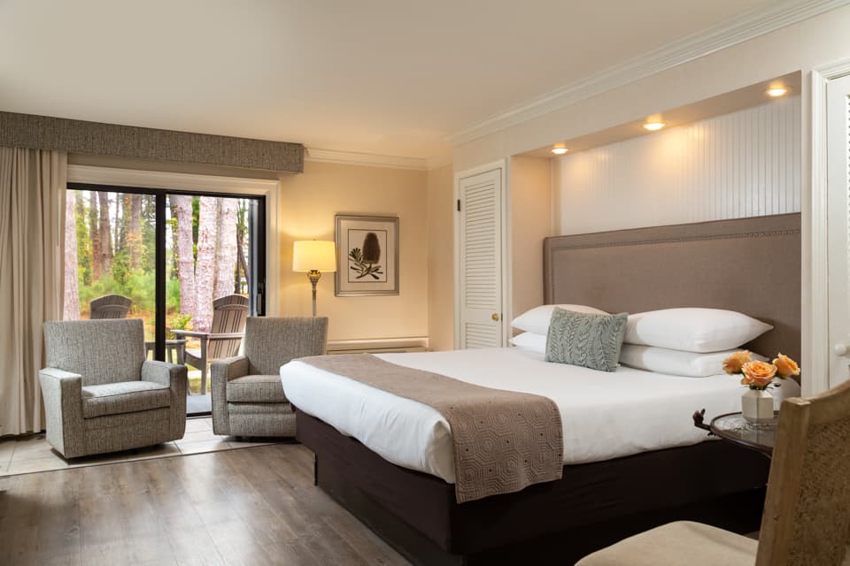 A cozy hotel room featuring a king-sized bed, two armchairs, and sliding doors leading to a terrace with outdoor seating.