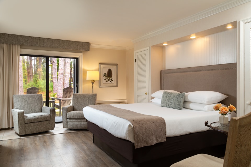 A cozy hotel room featuring a king-sized bed, two armchairs, and sliding doors leading to a terrace with outdoor seating.