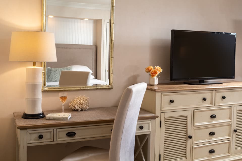 A cozy hotel room featuring a desk, a lamp, a TV, and decorative flowers.