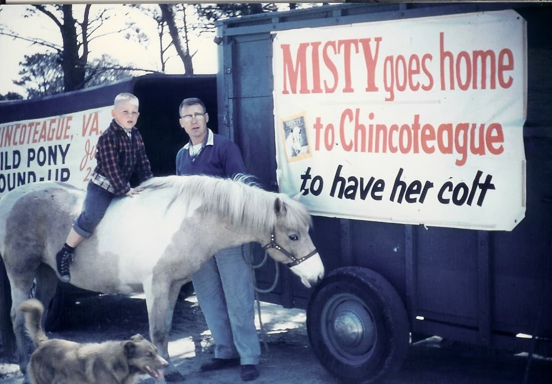 A boy sits on a white pony beside a man and a dog, with a sign that reads "MISTY goes home to Chincoteague to have her colt."