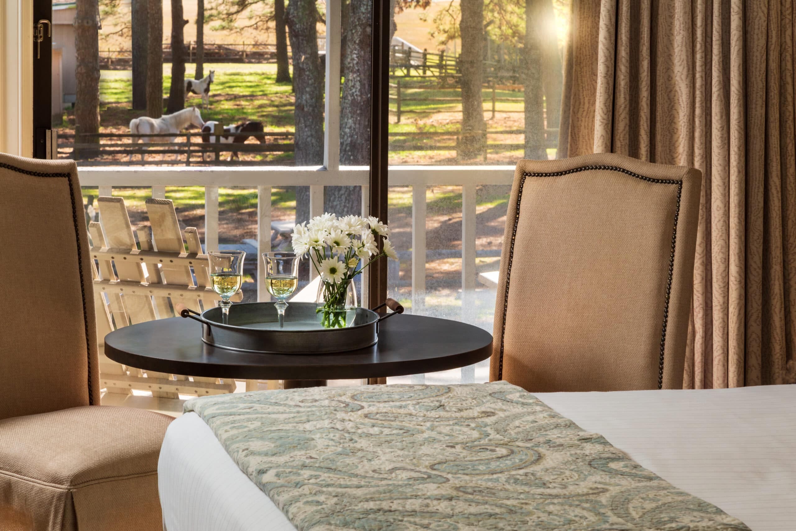 A cozy hotel room with two chairs, a table holding a floral arrangement and glasses, and a view of horses outside.