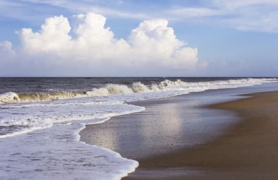 A sandy beach shoreline with gentle waves and fluffy clouds in the sky.