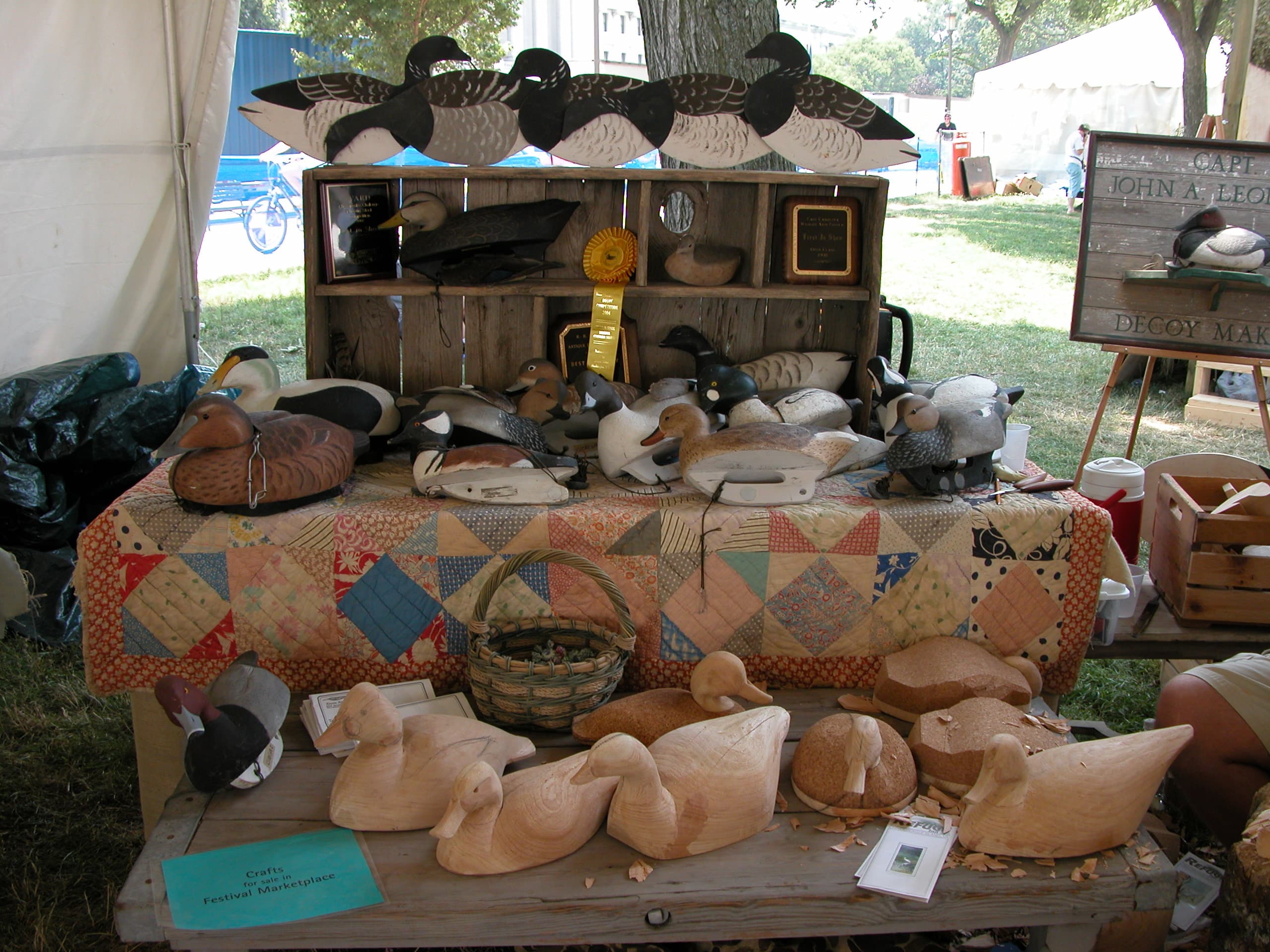 Hand-carved decoys at every stage of the work: roughed-out cedar bodies in the front row, primed and sanded birds behind them, and finished painted birds above, beside the Capt. John A. Leonard Decoy Maker sign.