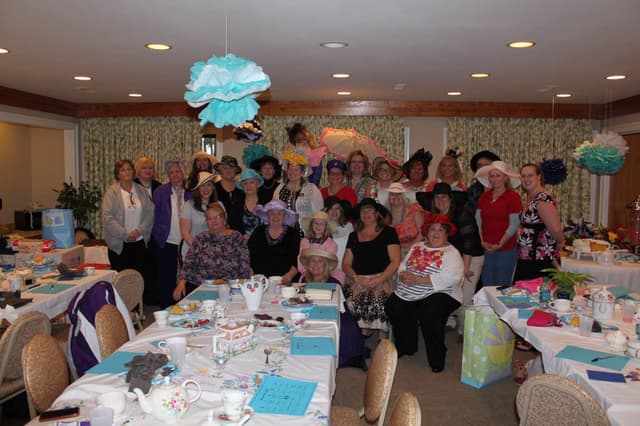 A large group in decorated hats gathered around the party tables in the Refuge Inn meeting room.