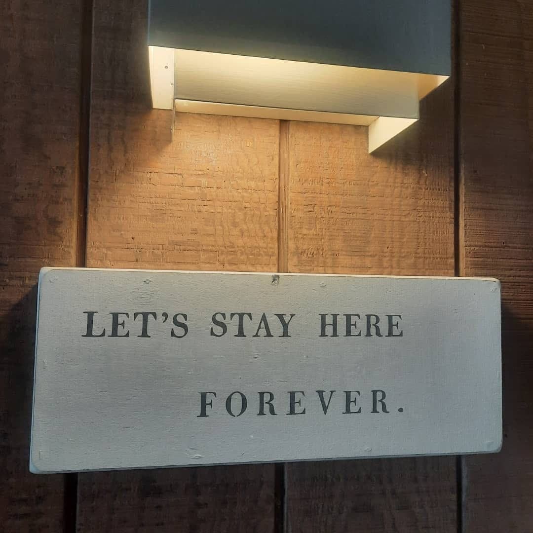 A wooden sign on a wall reads, "LET'S STAY HERE FOREVER" illuminated by a light above.