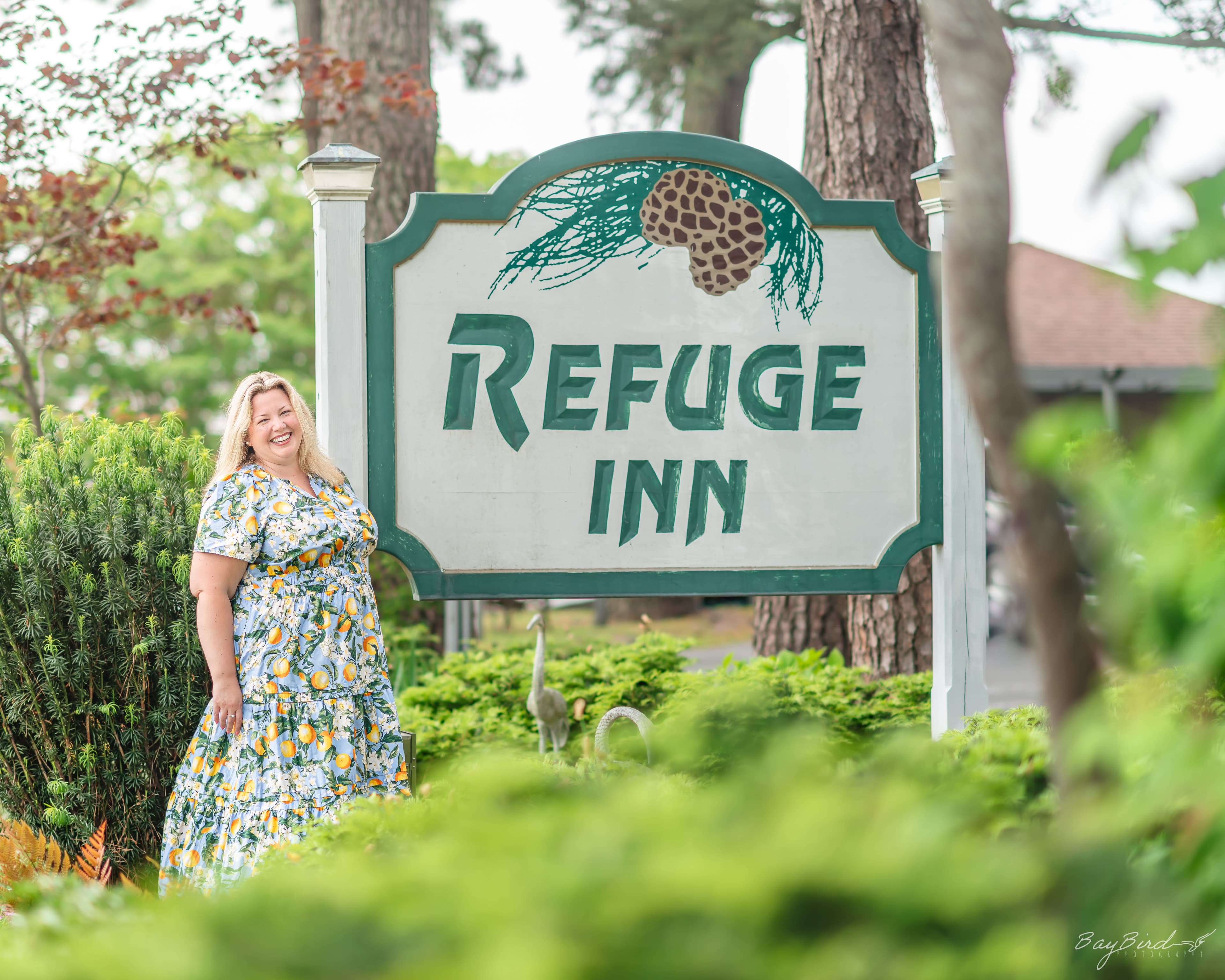 A smiling woman in a floral dress stands next to a sign for the Refuge Inn surrounded by greenery.