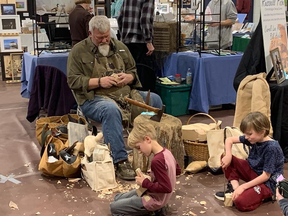 A bearded man carves wood while two young children sit on the ground beside him, surrounded by shavings and tools. A bearded man carves wood while two young children sit on the ground beside him, surrounded by shavings and tools.
