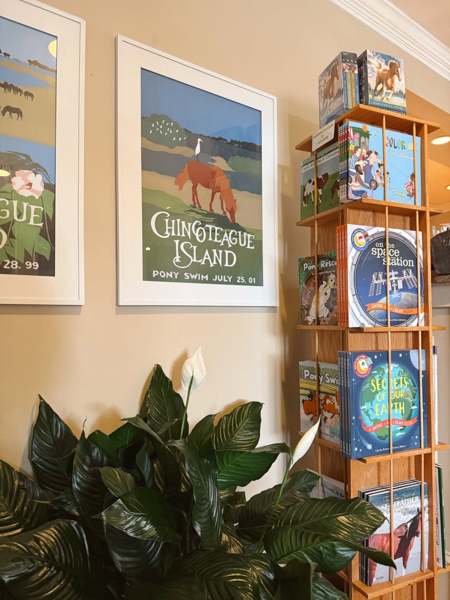 A corner of a room featuring framed art of Chincoteague Island and a wooden bookshelf filled with colorful children's books.