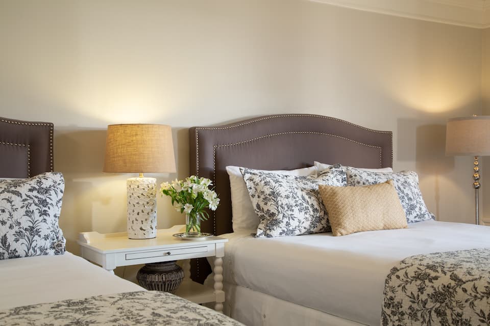 A cozy bedroom featuring two neatly made beds, decorative pillows, stylish lamps, and a small vase of flowers on a bedside table.