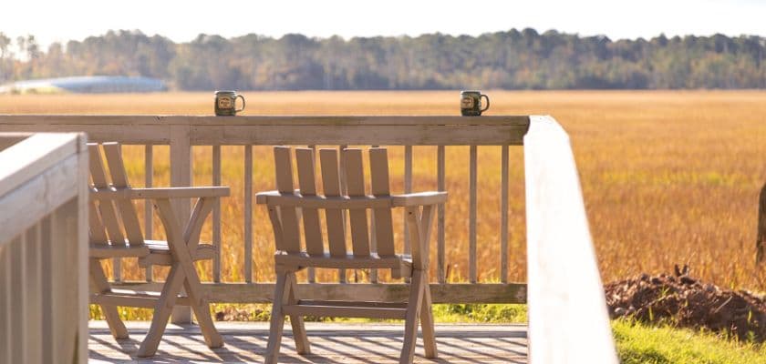 Two wooden chairs on a deck overlook a golden field with two mugs resting on the railing.