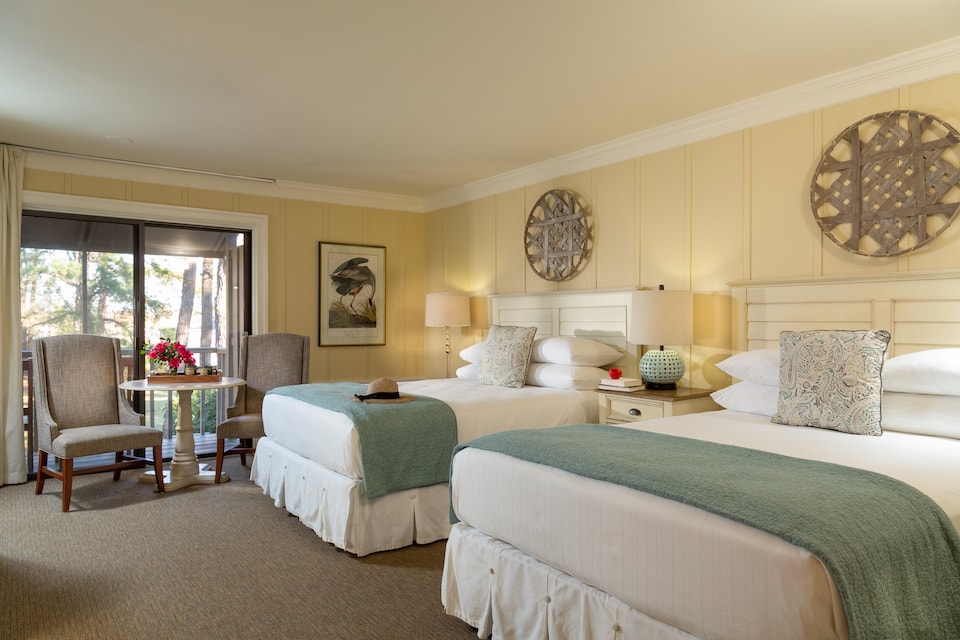A cozy hotel room featuring two beds, a small table with chairs, and a view of trees through a sliding door.