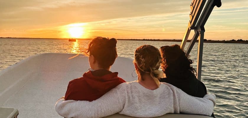 Three people sit side by side on a boat at sunset, facing the water with the sun low on the horizon