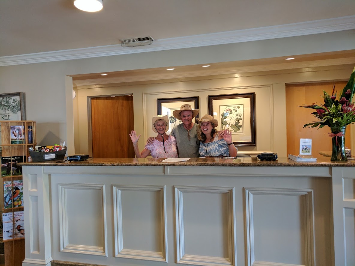 3 staff members in cowboy hats stand behind the Refuge Inn front desk, waving and smiling.