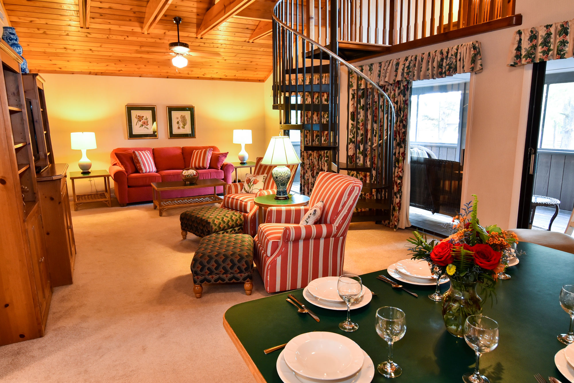 A cozy room with cream carpet and wooden cathedral ceilings. There are two armchairs and a red sleeper sofa. There is a spiral staircase leading to a loft, and several sliding doors leading to a private screened deck.